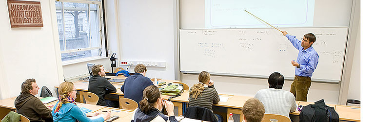 Ein Vortragender erklärt an der Tafel vor einer Gruppe Studierender.