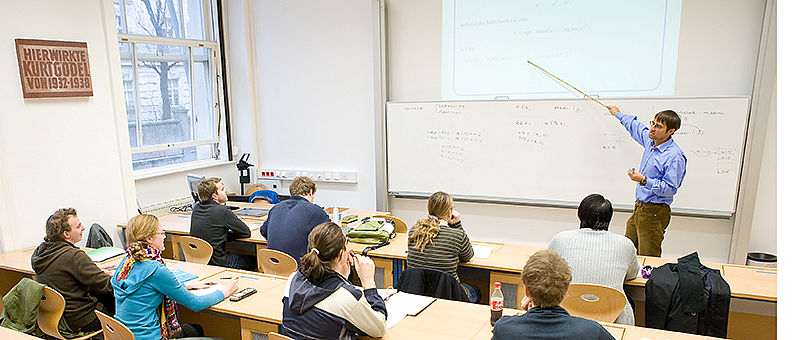 Ein Vortragender erklärt an der Tafel vor einer Gruppe Studierender.